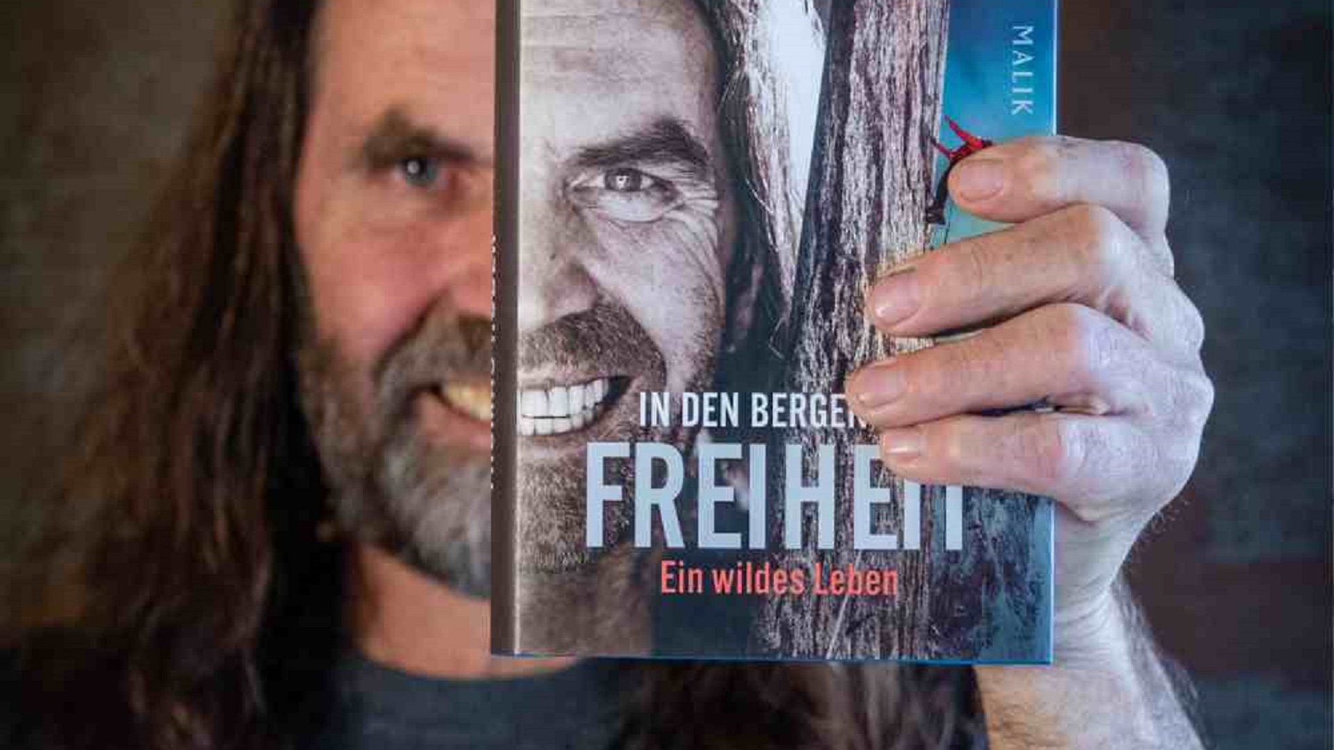 Foto für die Veranstaltung "Multivisionsvortrag: In den Bergen ist Freiheit" mit dem Künstler, der die Hälfte seines Gesichts zeigt, während er ein Bild in einem Buch hält.