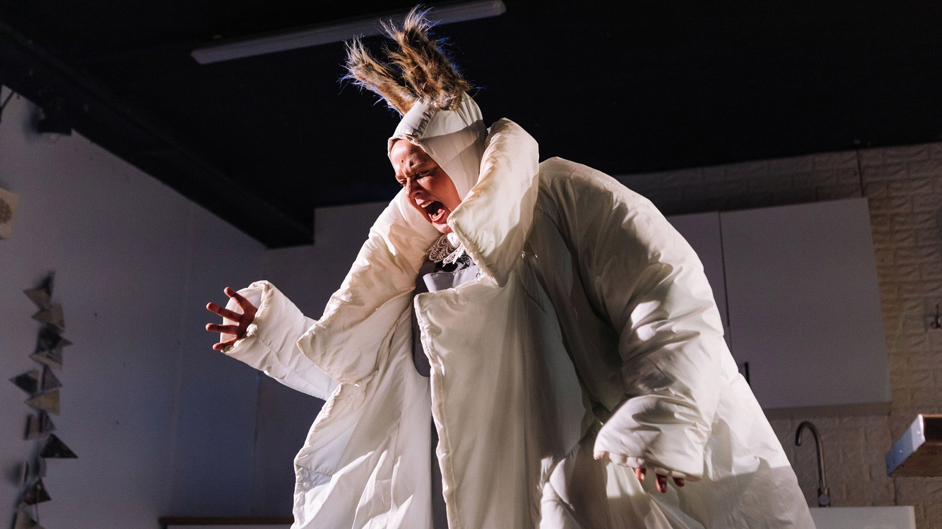 Person auf einer Theaterbühne in einem voluminösen weißen Kostüm mit auffälligem Kopfschmuck. Die Person steht im Mittelpunkt der Szene und wird von warmem Bühnenlicht beleuchtet. Im Hintergrund sind Teile eines Bühnenbilds mit hellen Wänden und Einrichtungsdetails zu erkennen.