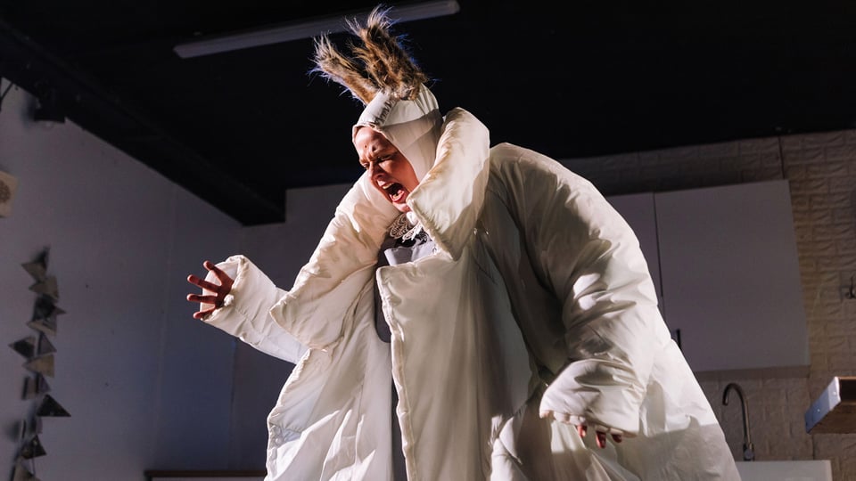 Person auf einer Theaterbühne in einem voluminösen weißen Kostüm mit auffälligem Kopfschmuck. Die Person steht im Mittelpunkt der Szene und wird von warmem Bühnenlicht beleuchtet. Im Hintergrund sind Teile eines Bühnenbilds mit hellen Wänden und Einrichtungsdetails zu erkennen.