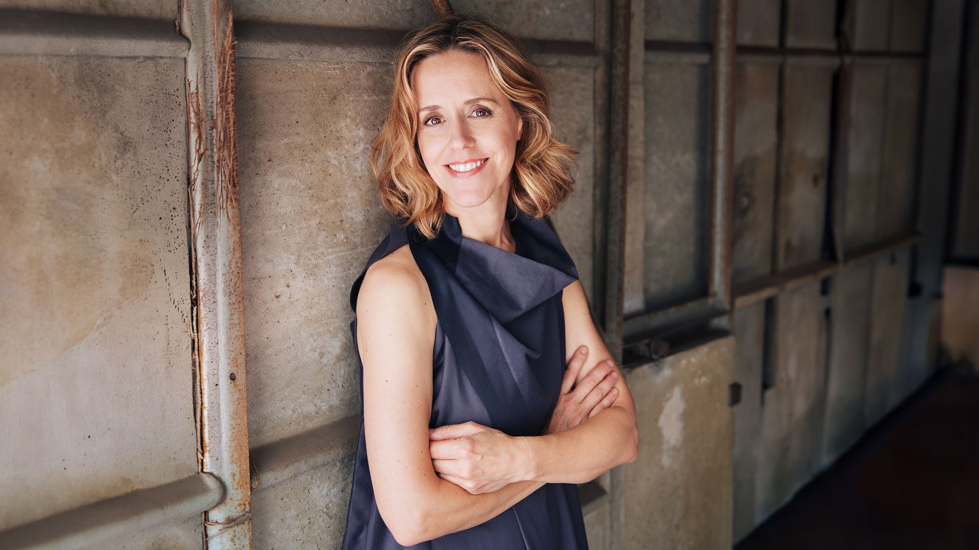 Porträtmotiv von Sol Gabetta: Die Cellistin steht mit verschränkten Armen vor einer industriell wirkenden Wand aus Metallplatten. Sie trägt ein ärmelloses, dunkles Kleid. Das Bild ist seitlich aufgenommen, mit weichem, natürlichem Licht von links, das Gesicht und Oberkörper gleichmäßig ausleuchtet. Ruhiges, hochwertiges Promofoto zur Ankündigung eines klassischen Orchester‑ und Cellokonzerts mit Les Siècles.