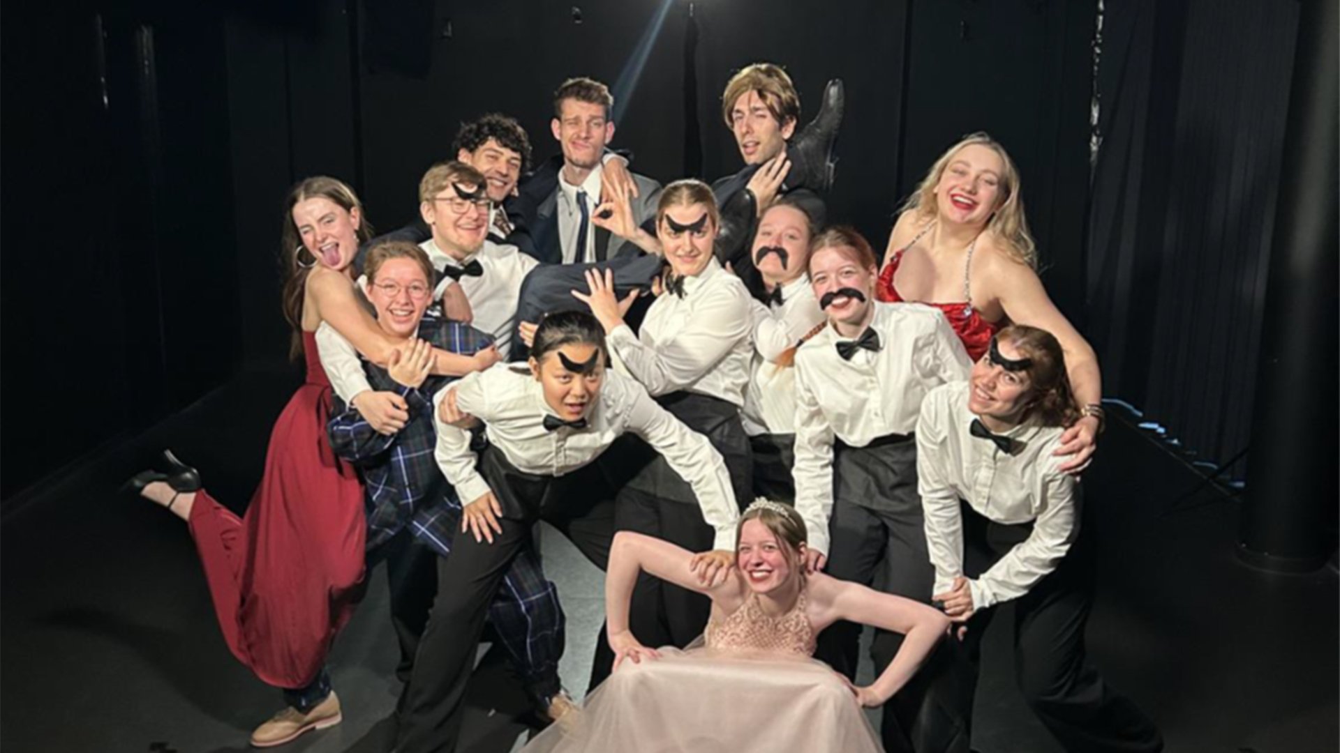 Gruppenfoto aus Mr. Marmalade mit zwölf Darstellenden in Abendgarderobe und weißen Hemden mit Fliegen oder aufgeklebten Schnurrbärten, die in einer engen Formation auf einer dunklen Bühne posieren, einige tragen andere Ensemblemitglieder oder werden von ihnen gestützt