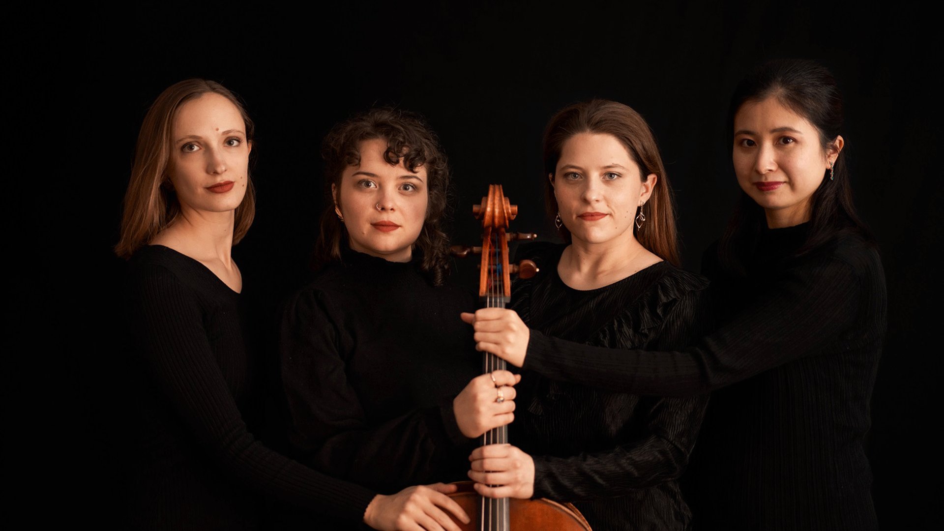 Porträt des Moser String Quartets vor schwarzem Hintergrund. Vier Musikerinnen in schwarzer Konzertkleidung stehen eng beieinander und halten gemeinsam ein Violoncello in der Bildmitte.
