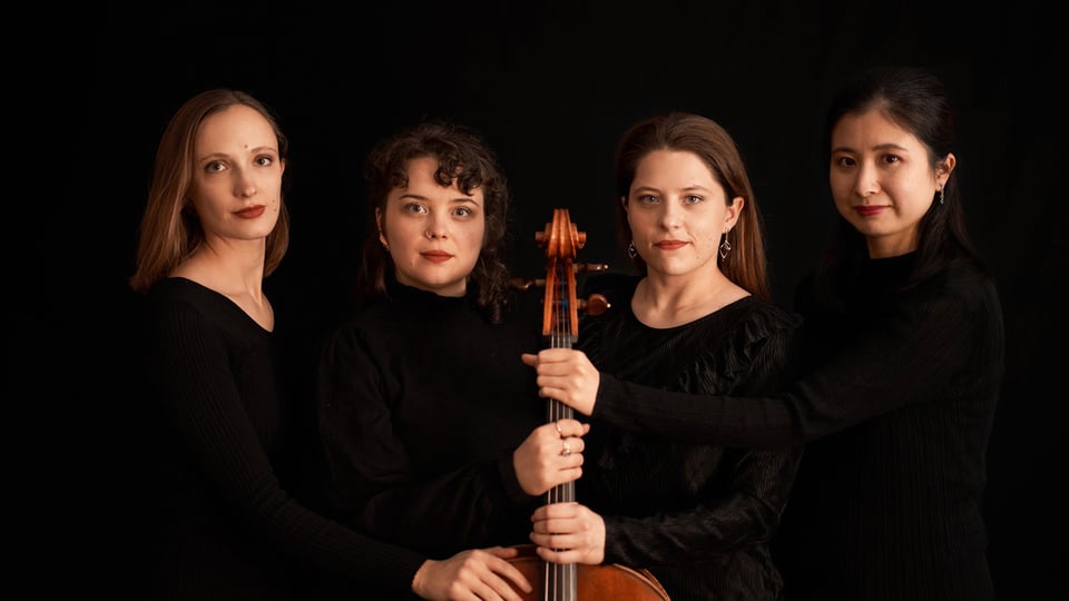 Porträt des Moser String Quartets vor schwarzem Hintergrund. Vier Musikerinnen in schwarzer Konzertkleidung stehen eng beieinander und halten gemeinsam ein Violoncello in der Bildmitte.