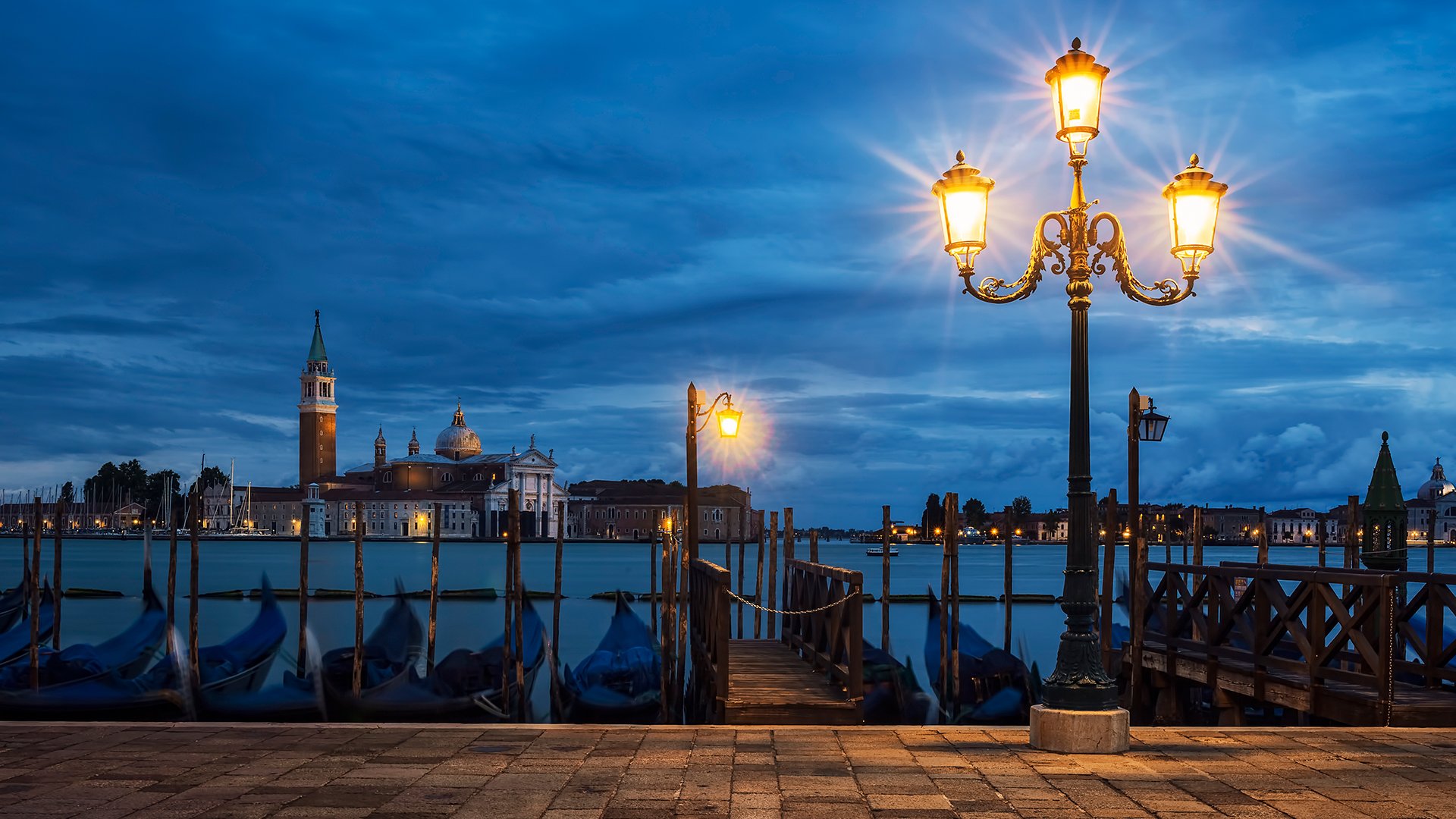 Abendliche Szene am Wasser in Venedig mit beleuchteten Laternen, Gondeln im Vordergrund und historischer Kirche auf gegenüberliegender Uferseite unter blauem Himmel.