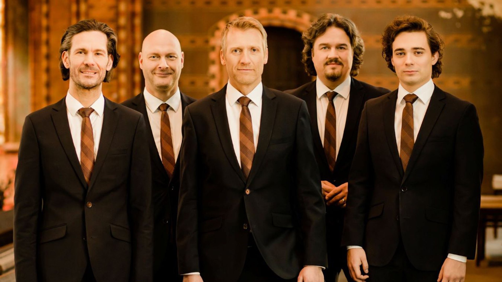 Fünf Männer in schwarzen Anzügen mit Krawatten stehen nebeneinander in einem festlichen Innenraum und blicken zur Kamera