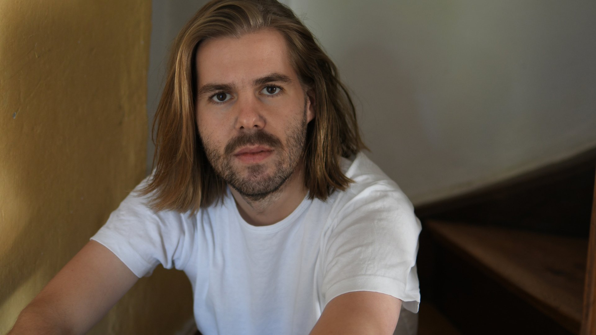 Porträtfoto von Philipp Schiepek in weißem T Shirt, mit schulterlangen Haaren und Bart, sitzend auf einer Holztreppe vor hellem Hintergrund und mit direktem Blick zur Kamera.