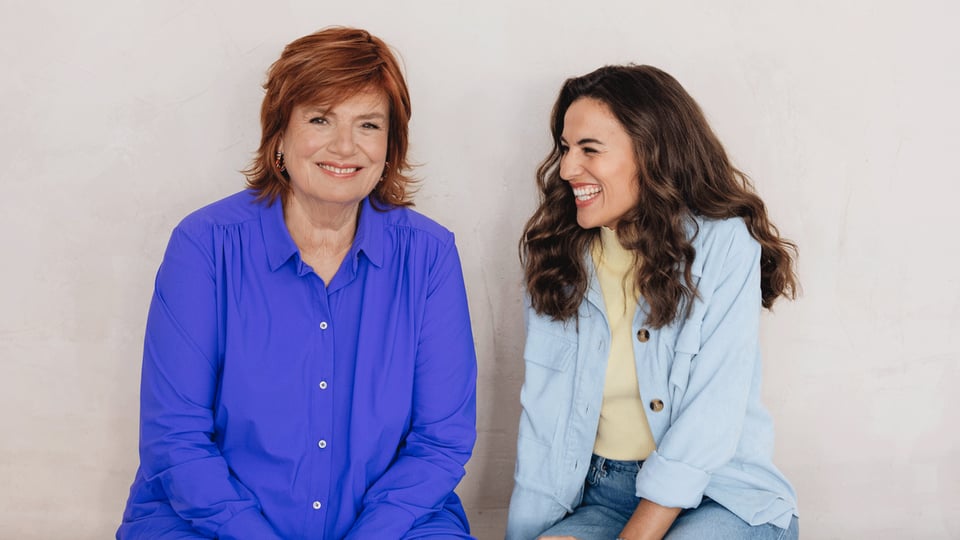 Portrait von Mona Ameziane und Christine Westermann vor hellem Hintergrund. Eine Person mit rotem Haar trägt eine blaue Bluse und blickt in die Kamera, während die andere Person in heller Jacke und Jeans seitlich zu der ersten Person gewandt ist.
