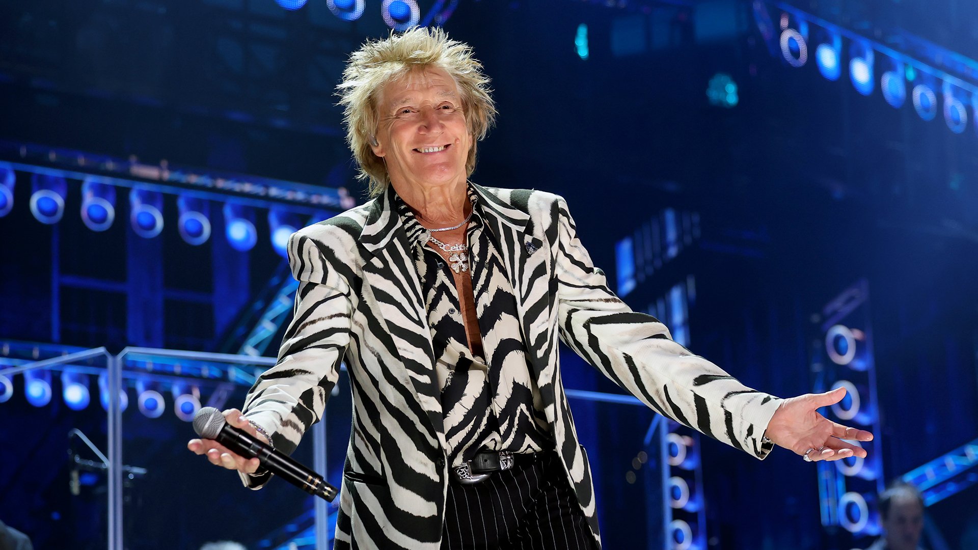 Bühnenfoto von Rod Stewart in schwarz weiß gemustertem Sakko mit Mikrofon in der Hand auf einer blau beleuchteten Konzertbühne, umgeben von Scheinwerfern und Bühnenelementen im Hintergrund