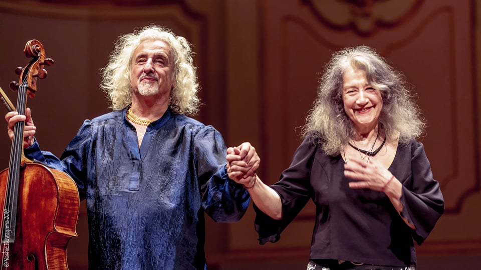 Zwei Musiker*innen stehen auf einer Konzertbühne, halten sich an den Händen; links eine Person mit Cello in dunkler Bluse, rechts eine Person in schwarzem Oberteil, vor warm ausgeleuchtetem Bühnenhintergrund.