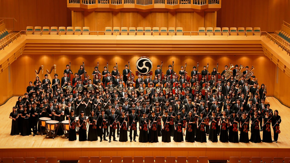 Großes Orchester des Waseda Symphony Orchestra Tokyo auf einer Konzertbühne mit zahlreichen Musiker:innen in schwarzer Kleidung und verschiedenen Instrumenten.