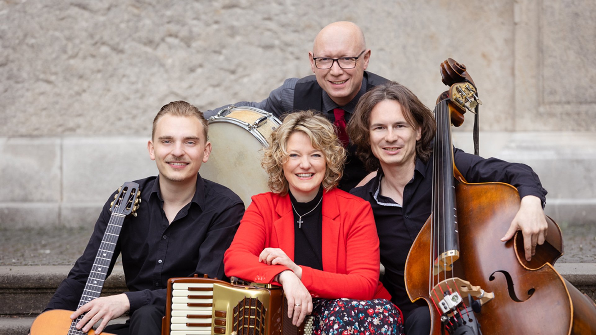 Drei Herren und eine Dame mit Instrumenten vor einer historischen Mauer sitzend 