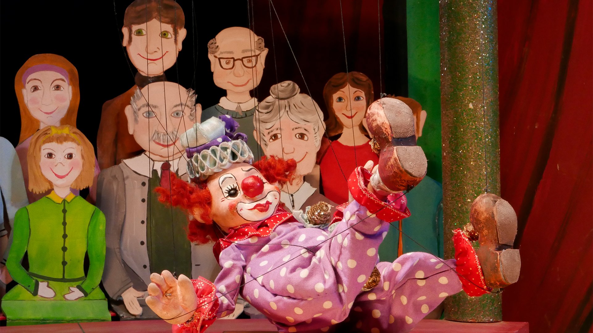 Eine bunte Clown-Marionette in lila gepunktetem Kostüm liegt spielerisch auf einer kleinen Bühne; im Hintergrund sind weitere Puppenfiguren einer Zuschauerreihe vor einem dekorierten Bühnenbild zu sehen.