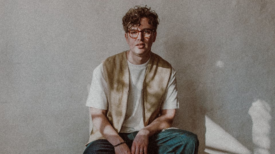 Paul Siemers mit runder Brille, bekleidet mit beigem Shirt und Weste sowie dunkler Hose und weißen Sneakern sitzt auf einem kleinen Koffer vor heller Wand