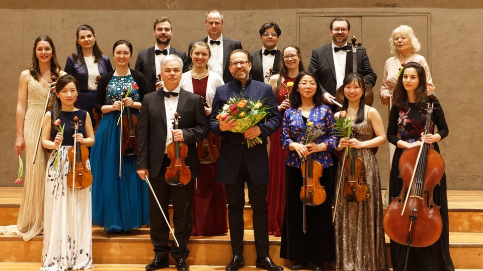 Ein Gruppenfoto des festlich gekleidetetn orchesters mit Instrumenten sowie einer Person ind er Mitte mit Blumenstrauß