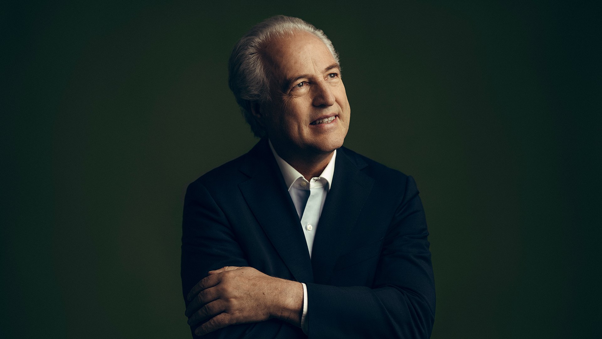 Manfred Honeck im dunklen Anzug und weißem Hemd, vor einem weich ausgeleuchteten grünlichen Hintergrund, mit verschränkten Armen und leicht zur Seite gewandtem Blick.