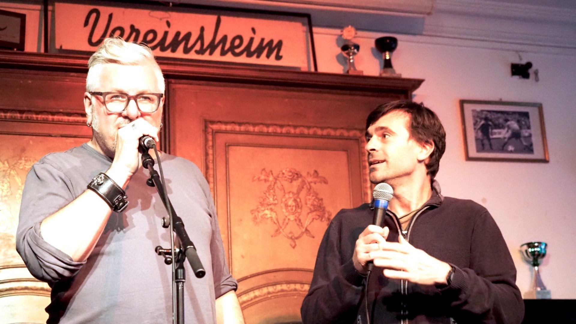 Bühnenfoto mit zwei Personen, die Mikrofone halten und auf einer kleinen Bühne sprechen, vor einer holzverkleideten Wand mit dem Schriftzug Vereinsheim sowie gerahmten Bildern und Pokalen im Hintergrund.