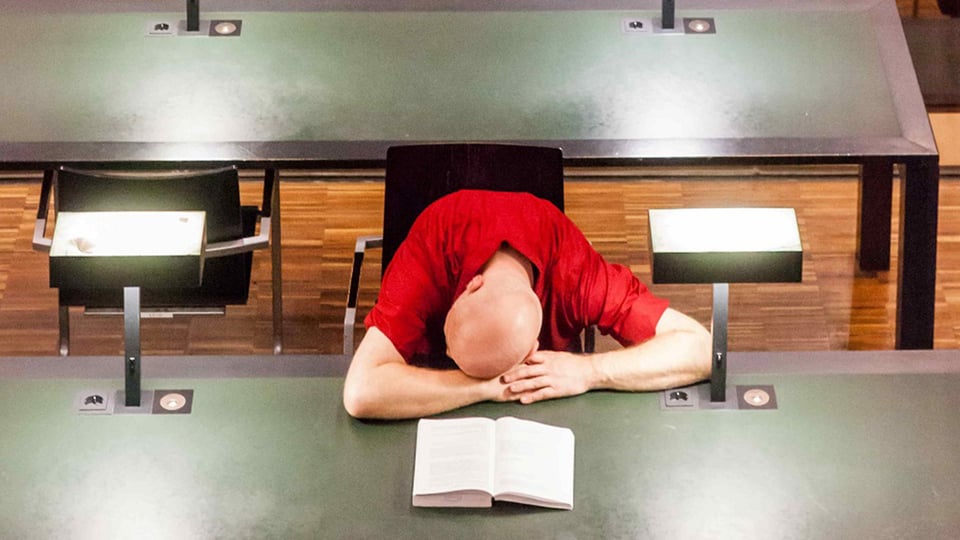 Foto für die Veranstaltung "Horst Evers – So gesehen natürlich lustig" mit einem Mann, der an einem grünen Tisch sitzt, ein geöffnetes Buch hält und es in seinen Armen auf dem Tisch liegend betrachtet.