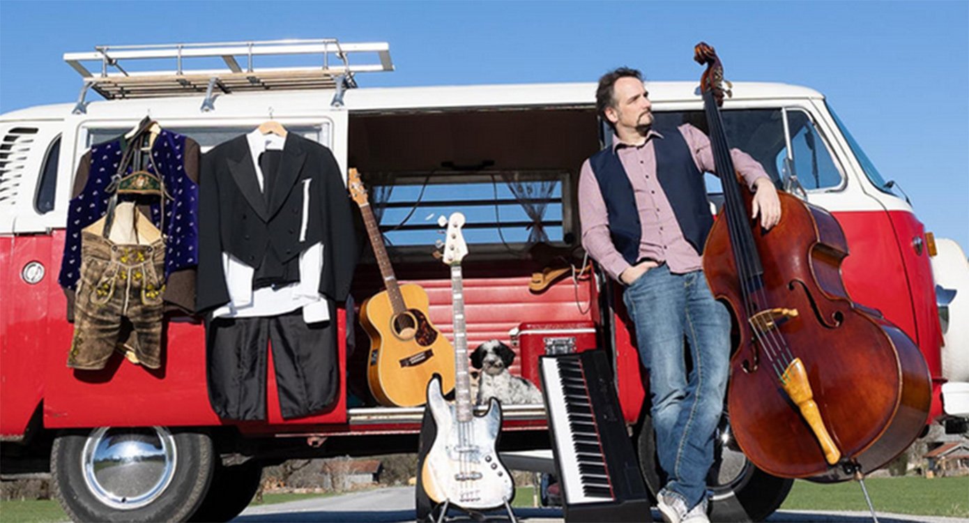 Mann mit Cello in Freizeitkleidung vor einem VW Bus stehend mit weiteren Instrumenten und aufgehängter Kleidung
