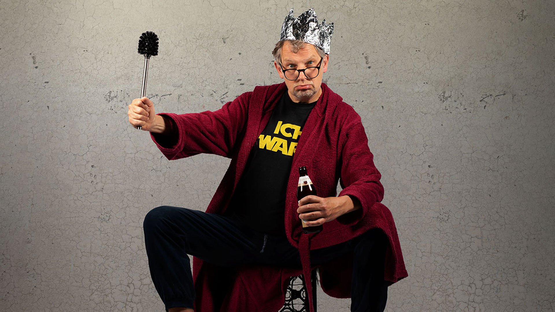 Foto für die Veranstaltung von Severin Groebner – ICH BIN DAS VOLK mit dem Künstler, der auf einem Stuhl sitzt, in einer Hand ein Bier hält und in der anderen eine Toilettenbürste, während er eine Krone aus Alufolie trägt; im Hintergrund eine Steinwand