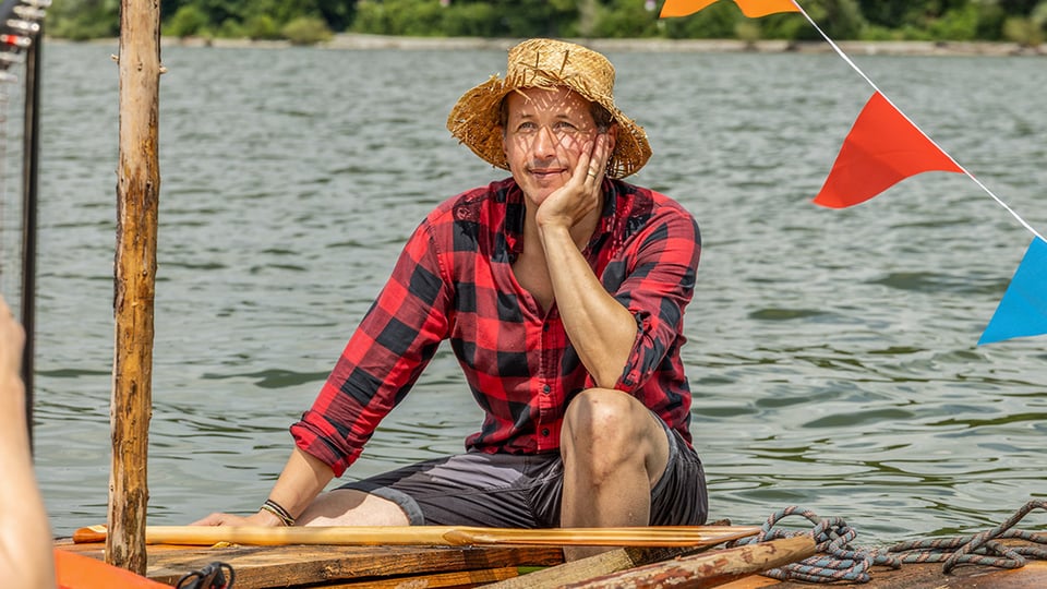 Foto von Willi Weitzel auf einem Holzfloß auf einem See, mit Strohhut, rot schwarz kariertem Hemd und Shorts, daneben bunte Wimpel, Seile und Holzstangen vor bewaldetem Ufer im Hintergrund.