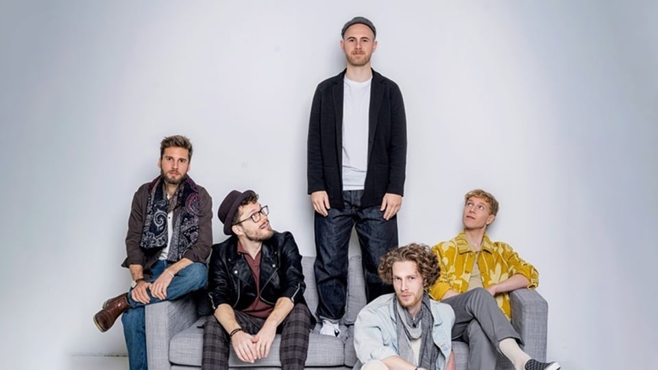 Die fünfköpfige Band auf einem grauen Sofa mit grauem Hintergrund