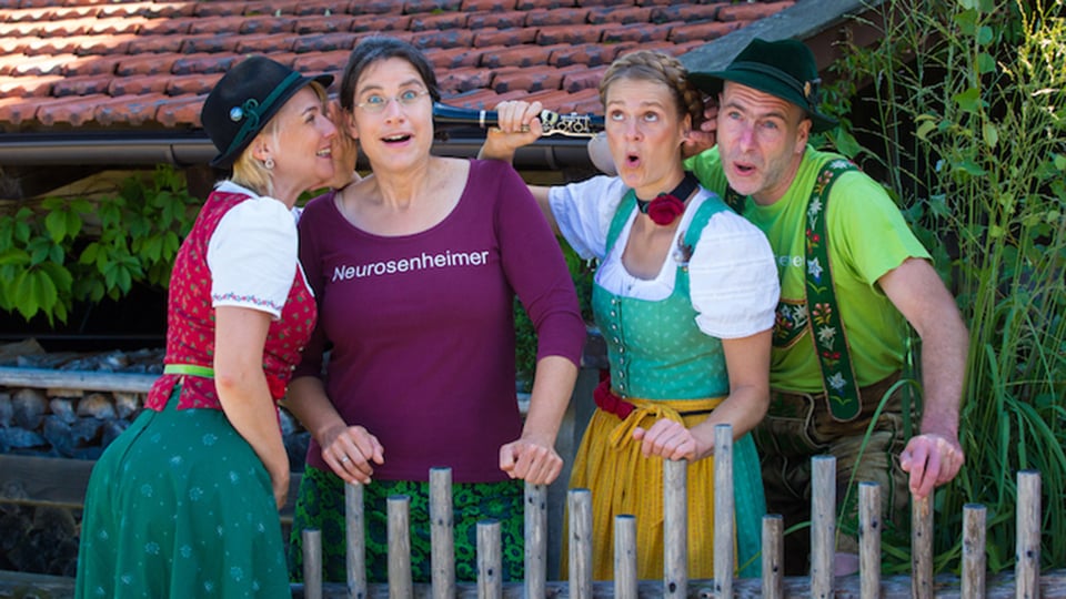 Vier Personen stehen hinter einem niedrigen Holzzaun, drei tragen traditionelle Tracht mit Hüten und eine Person ein bordeauxfarbenes T‑Shirt mit der Aufschrift Neurosenheimer, im Hintergrund ein Haus mit Ziegeldach und Garten.