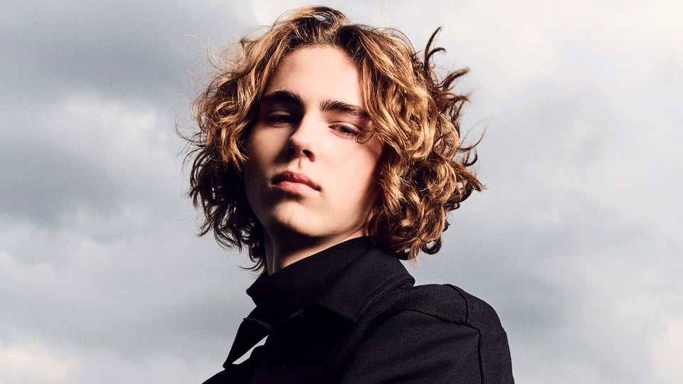 Porträt von Justus Eichhorn mit lockigem schulterlangem Haar und schwarzem Mantel vor bewölktem Himmel. Die Aufnahme zeigt ihn aus leichter Untersicht mit Blick zur Kamera.