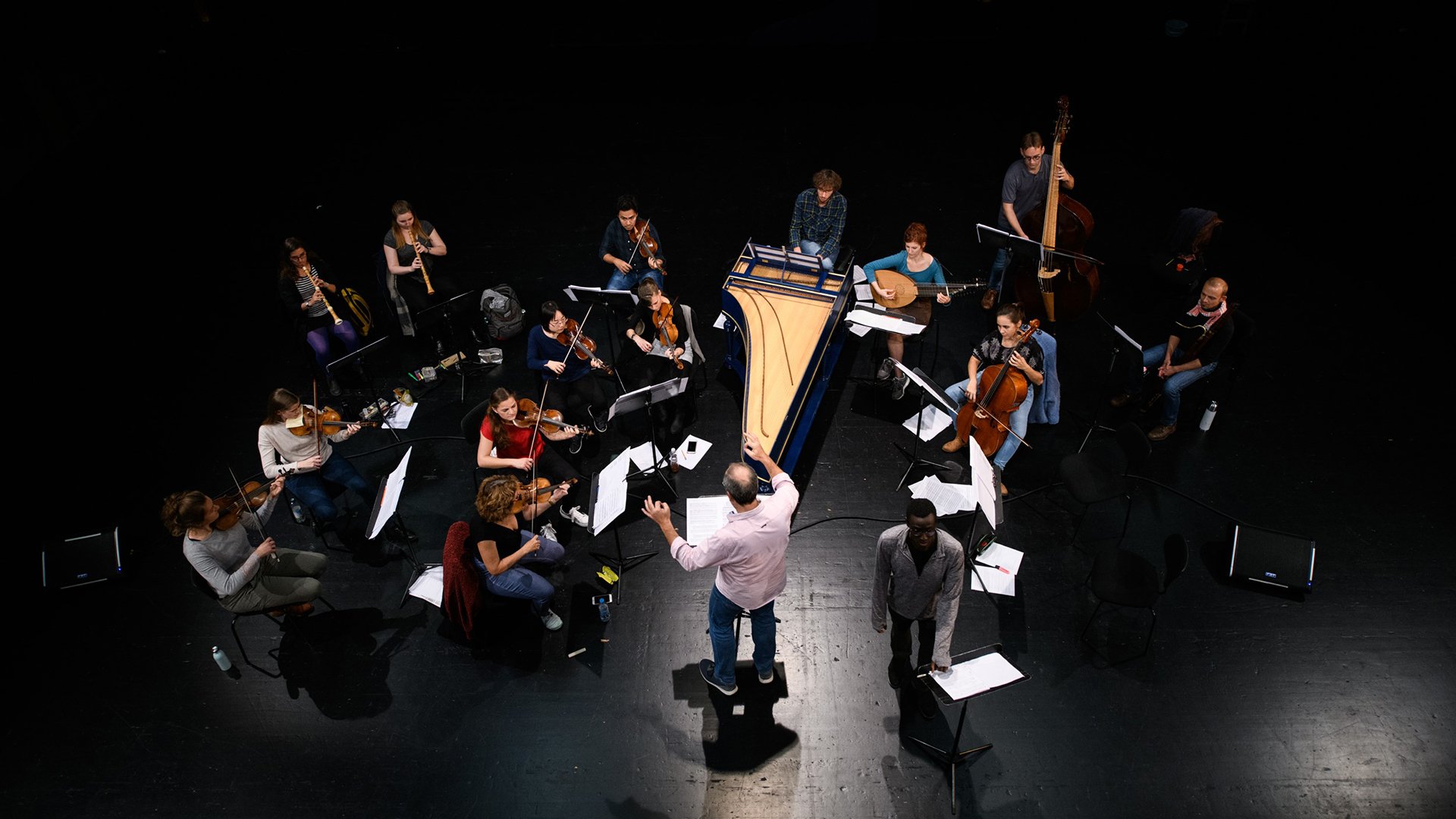 Bild eines Orchesters aus der Vogelperspektive mit Instrumenten auf einem schwarzen Bühnenboden stehend