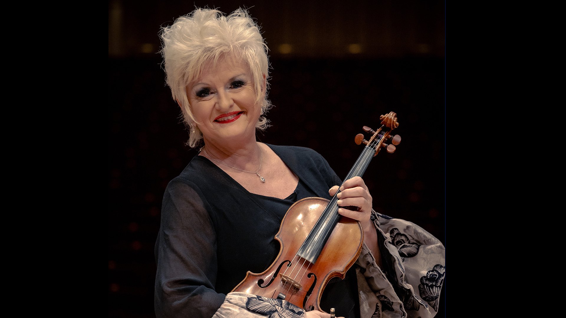 Foto der Violinistin Ilona Cudek in festlicher Kleidung mit Instrument vor dunklem Hintergrund