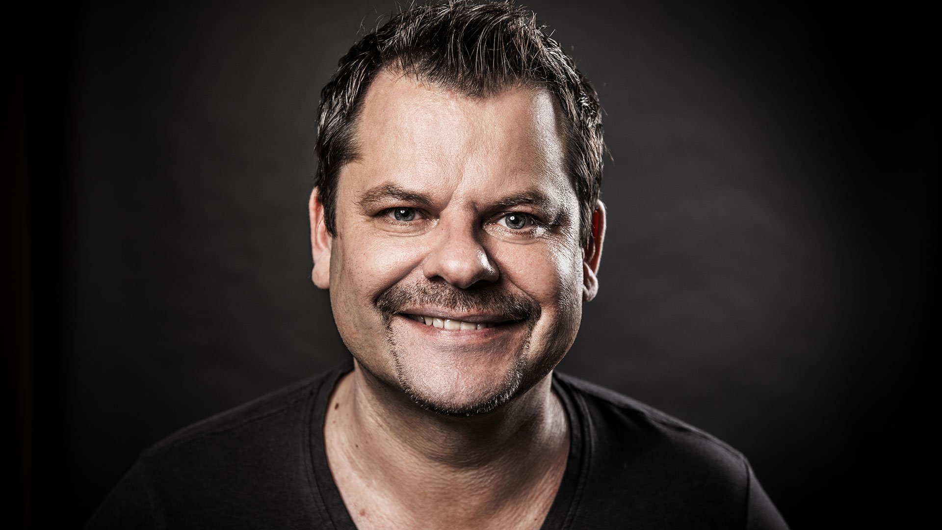 Porträtfoto von Ingo Appelt vor dunklem Hintergrund, mit kurzem Haar und schwarzem T-Shirt, lächelnd in die Kamera.