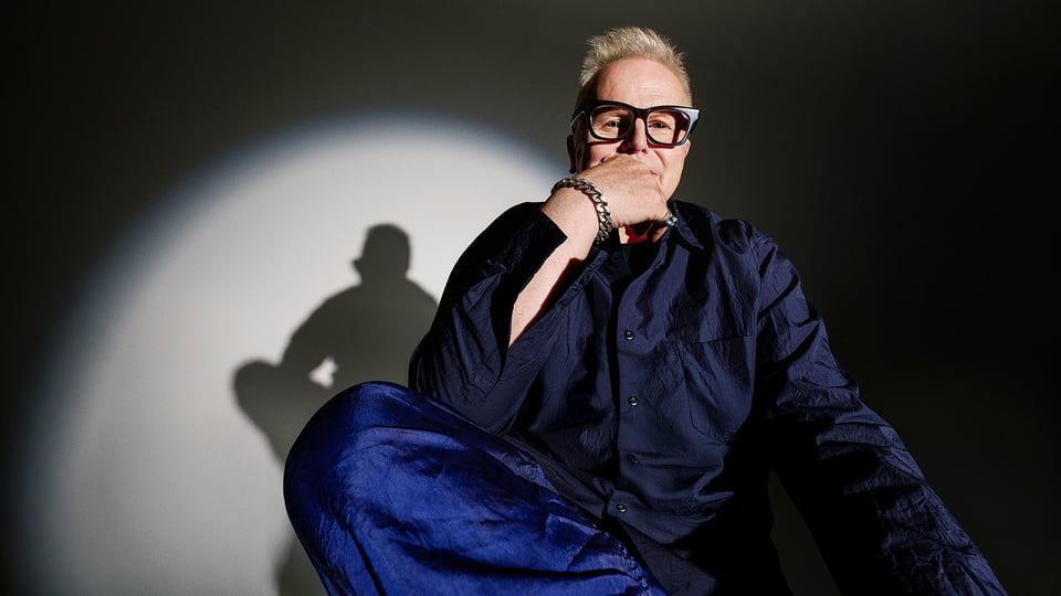 Herbert Grönemeyer sitzt vor einem grauen Hintergrund im Scheinwerferlicht und hat ein blaues Outfit an