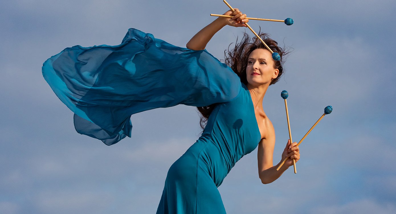 eine Dame in blauem Kleid mit Klöppeln in der Hand vor helllblauem Hintergrund